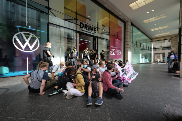 Eine Gruppe von Menschen sitzt auf dem Boden vor einer Volkswagen-Vertretung mit Glaswänden und Logo, die eine Fahne mit Text halten.