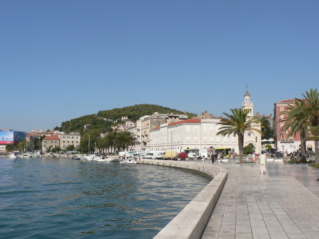 Boote auf dem Wasser links, Menschen auf der Straße rechts, mit Fahrzeugen, Bäumen, Gebäuden, einem Berg und einem blauen Himmel im Hintergrund.