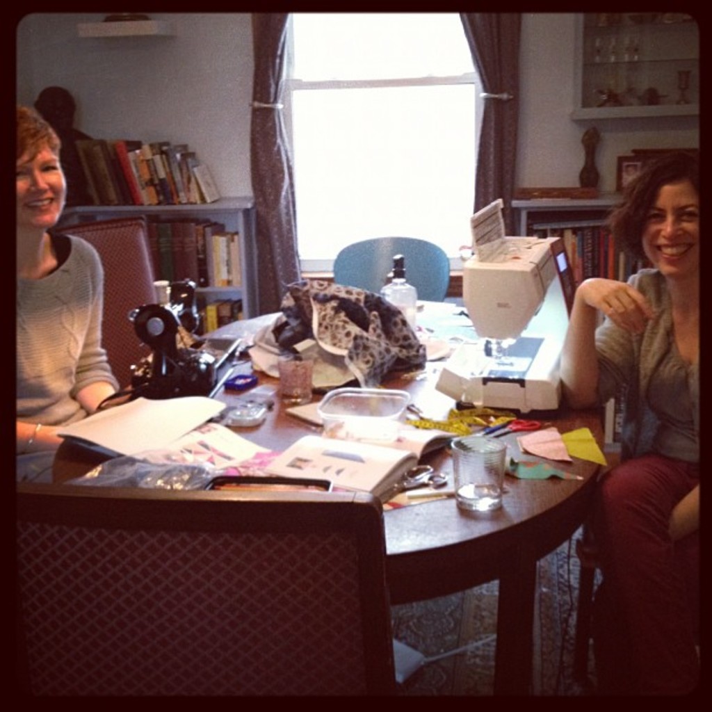 Zwei Frauen an einem Tisch mit einer Nähmaschine, Papieren und einem Glas, mit einem Bücherregal und Vorhängen im Hintergrund.