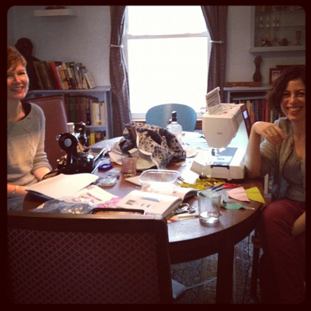 Zwei Frauen an einem Tisch mit einer Nähmaschine, Papieren und einem Glas, mit einem Bücherregal und Vorhängen im Hintergrund.