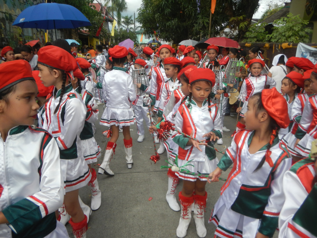 Gruppe junger Mädchen in roten und weißen Uniformen marschiert mit Musikinstrumenten und Schirmen die Straße entlang, mit Bäumen, Gebäuden und einem klaren blauen Himmel im Hintergrund.