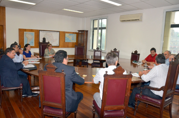 Eine Gruppe von Menschen, die an einem Tisch sitzen und Papiere und Gläser haben, ein Klimagerät, eine Mitteilungstafel und Karten an der Wand im Hintergrund.