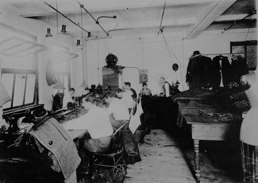 Ein Schwarz-Weiß-Bild von Menschen, die auf Stühlen sitzen und Nähmaschinen verwenden, sowie ein Tisch mit Kleidung auf der rechten Seite und Wände mit Fenstern auf der linken Seite und verschiedenen Gegenständen im Hintergrund.
