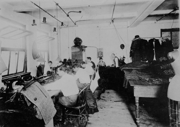 Ein Schwarz-Weiß-Bild von Menschen, die auf Stühlen sitzen und Nähmaschinen verwenden, sowie ein Tisch mit Kleidung auf der rechten Seite und Wände mit Fenstern auf der linken Seite und verschiedenen Gegenständen im Hintergrund.