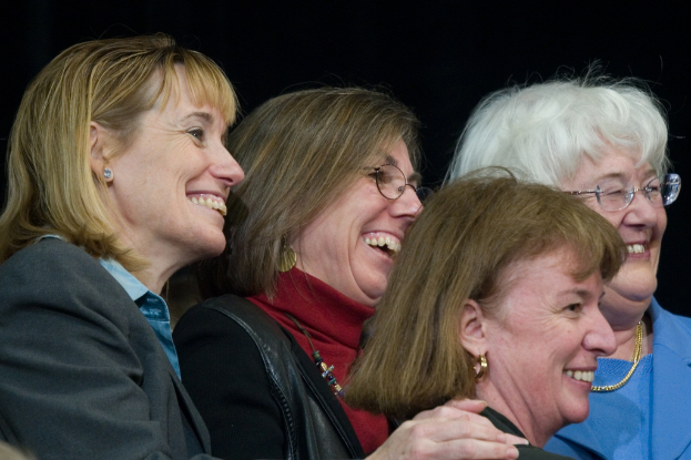 Mehrere ältere Frauen stehen in der Vordergrund, lächeln und schauen nach rechts, mit einem schwarzen Hintergrund dahinter.
