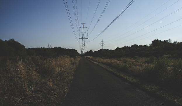 Straße mit Stromleitungen im Freien, umgeben von grünem Gras und Bäumen, die zu fernen Übertragungsmasten führt.