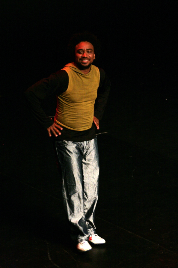 Eine Person in einem T-Shirt lächelt und performs auf einer Bühne mit einem dunklen Hintergrund.