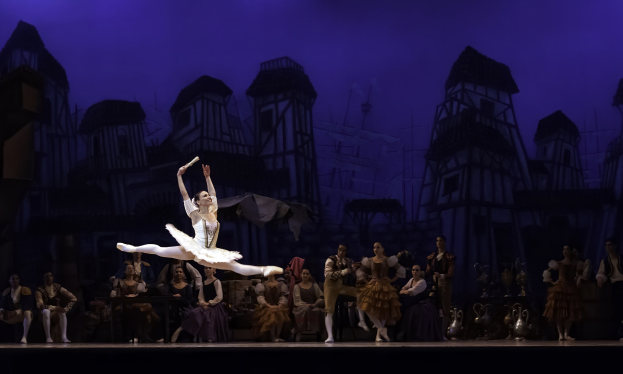 Eine Balletttänzerin in einem weißen Tutu und Spitzenschuhen tanzt auf der Bühne, während das Publikum zusieht, mit einer gemalten Burg als Hintergrund.
