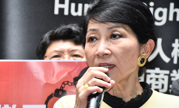 Eine Frau in einem schwarzen Kleid hält ein Mikrofon und steht selbstbewusst vor einer Menge mit einem Banner "Hongkong-Chief-Executive-Carrie-Lam" dahinter.