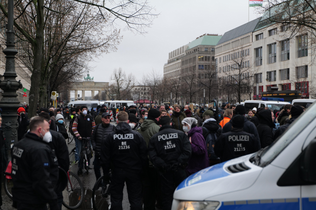Eine große Gruppe von Menschen steht vor einem Polizeiwagen auf einer Straße, die von Bäumen, Laternenmasten und Gebäuden mit Fenstern umgeben ist, einige tragen Mützen und Masken, mit Fahrrädern im Vordergrund und einem Bogen mit Säulen und einer Fahne im Hintergrund.