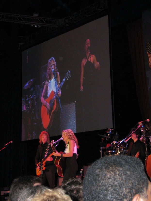 Ein Musikkonzert mit einem Publikum im Vordergrund, drei Musiker auf der Bühne: einer spielt Gitarre, einer singt und spielt Gitarre und einer spielt Schlagzeug, mit einem Projektor im Hintergrund.