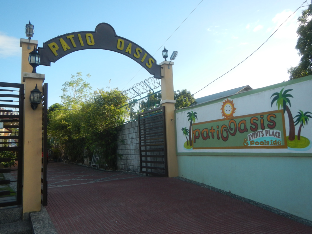 Eintritt zum Patio Oasis Events Place & Poolside mit einem Bogen mit Text, einer bemalten Wand, einem Tor, Wandlampen, Bäumen und einem bewölkten Himmel.