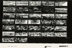 Schwarzes und weißes Foto einer vielseitigen Gruppe von Menschen, die um einen Fernseher versammelt sind und in die Kamera schauen, mit Text am unteren Rand.