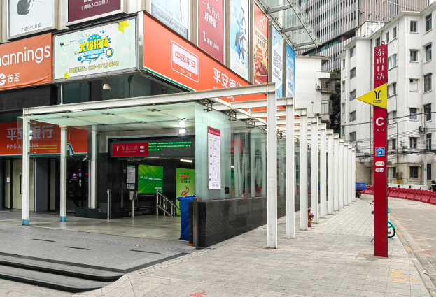 Eine Stadtstraße mit einem Bushaltestellen, Gebäude auf beiden Seiten, eine Straße mit einem Mülleimer in der Mitte, rote und gelbe Schilder mit Text auf der rechten Seite, Menschen auf dem Gehweg und eine Rolltreppe auf der linken Seite.