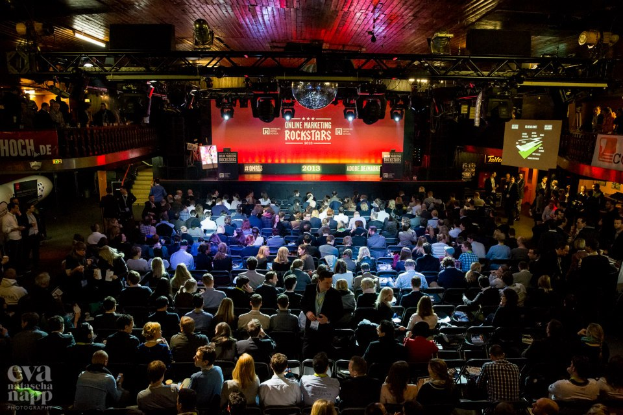 Eine große Menschenmenge sitzt auf Stühlen vor einer Bühne mit Bildschirmen und Bannern, beleuchtet von Scheinwerfern und umgeben von Geländern, mit einem Wasserzeichen in der linken unteren Ecke, das eine Atmosphäre von Spannung und Erwartung für ein Online-Marketing-Rockstars-Event schafft.