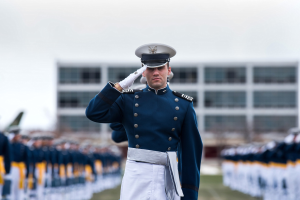 Ein Mann in militärischer Uniform salutiert auf einer Abschlussfeier umgeben von einer Gruppe von Menschen auf dem Boden, mit einem Gebäude und Himmel im Hintergrund.