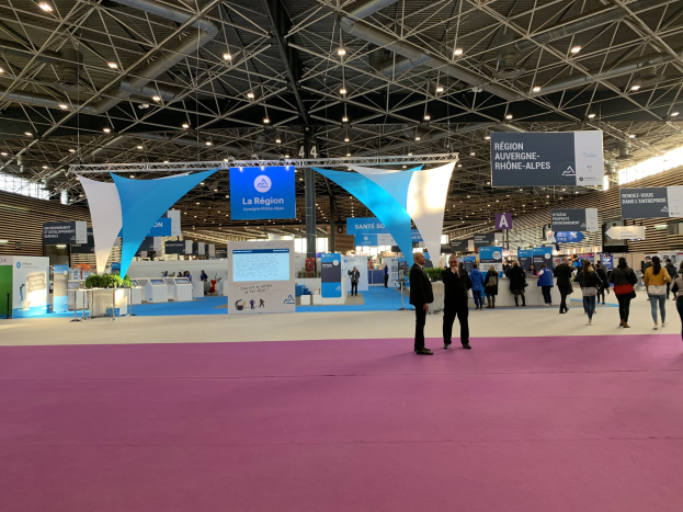 Ein großer Messestand auf der Internationalen Handelsmesse Paris 2018 mit Besuchern, die Taschen tragen und Tische mit Gegenständen unter beleuchteten Bannern betrachten.