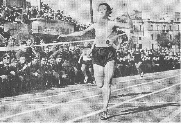 Ein Schwarz-Weiß-Foto einer Frau beim Laufen auf einer Bahn während des Frauen-100m-Finals bei den Olympischen Spielen 1956, mit einer Menschenmenge, Gebäuden und Pfählen im Hintergrund.