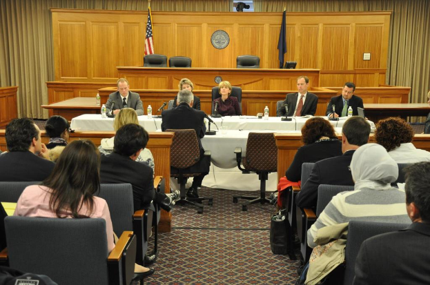 Ein Konferenzraum mit einer Gruppe von Menschen, die auf Stühlen sitzen, zwei ausländische Flaggen, ein oranger Schreibtisch mit Wasserflaschen und ein Mikrofon.