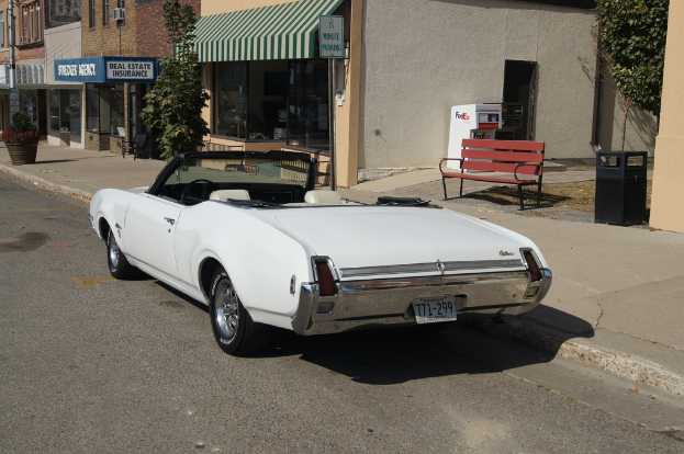 Ein weißes Auto mit der Nummer 771-299 steht auf einer Straße, rechts daneben eine Bank, im Hintergrund ein Gebäude und ein Geschäft namens 'Immobilienversicherung'.