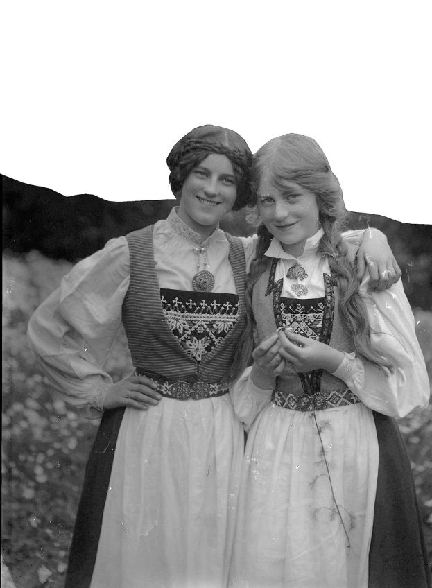 Zwei junge Frauen in traditioneller bayrischer Tracht, die weiße und schwarze Kleider mit aufwendiger Stickerei und Perlen tragen, posieren vor einer Kulisse aus grünen Pflanzen und einem hellen Himmel.