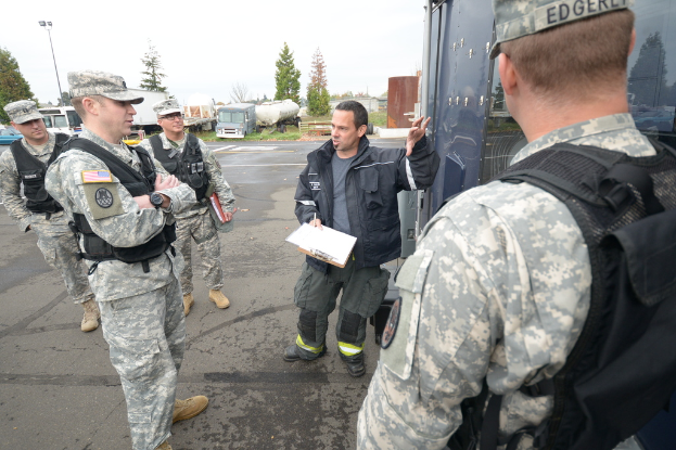 Eine Gruppe von Menschen in Uniformen, Mützen, Jacken und Schuhen steht auf einer Straße, wobei eine Person in einer schwarzen Jacke ein Klemmbrett mit Papieren und einen Stift in der Nähe eines Fahrzeugs hält; im Hintergrund sind weitere Fahrzeuge, Bäume, Gras, ein Zaun, ein Lichtmast und ein klarer Himmel zu sehen.