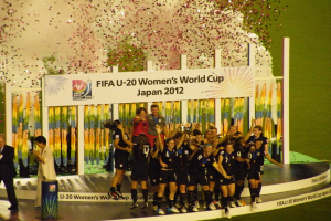 Schwarze-uniformierte Frauenfußballspielerinnen auf einer Bühne, die einen Sieg feiern, umgeben von einer Menge mit Kameras, mit einem Hintergrundboard und dekorativen Gegenständen auf Gras.