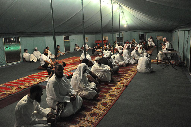 Eine Gruppe von Menschen sitzt auf dem Boden in einem Lager, wobei eine Person auf einem Stuhl rechts sitzt, ein Mikrofon vor ihnen, ein Tisch neben ihnen und eine Lampe oben im Bild.