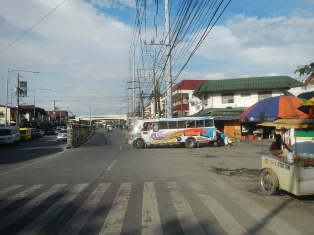 Eine Stadtstraße mit parkenden Fahrzeugen, darunter ein Bus, gesäumt von Gebäuden, Strommasten und Laternen, mit einer Brücke und einem bewölkten Himmel im Hintergrund.