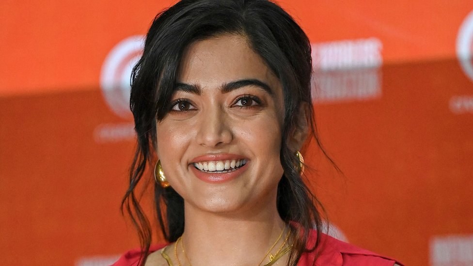Eine Frau in einem roten Kleid mit einem eleganten Dutt und dezentem Make-up lächelt in die Kamera, trägt schwarze High Heels und hat ein hellgelbes Banner mit der Aufschrift 'Die besten Schönheits Looks von Sonam Kapoor beim Cannes Film Festival' im Hintergrund.