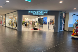 Das Innere eines Betty-Barclay-Ladens in einem Einkaufszentrum, das Schaufensterpuppen in Kleidern, hängende Kleidung, Tische, Pflanzen und ein Spielzeugfahrzeug auf der rechten Seite zeigt, mit einer Tafel oben, auf der "Betty Barclay Selected Brands" steht.