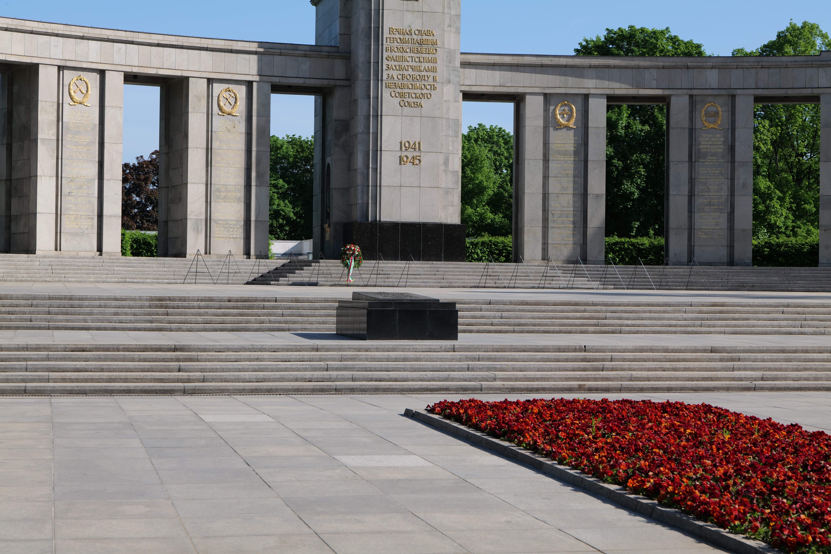 Ein Gedenkmonument in einem Park mit roten Blumen davor, mit Inschrift auf seinen Wänden und Stufen, umgeben von Bäumen unter einem klaren blauen Himmel.