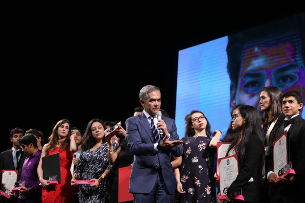 Ein Mann im Anzug und mit Krawatte hält ein Mikrofon auf einer Bühne, umgeben von Menschen mit Auszeichnungen, mit einem Bildschirm im Hintergrund.