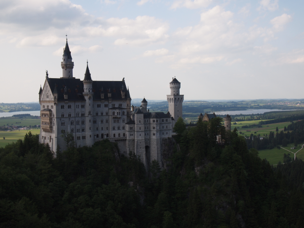 Neuschwanstein Burg in Deutschland, ein majestätisches Gebäude umgeben von saftig grünen Bäumen, einer gewundenen Straße, einem ruhigen Gewässer, rollenden Hügeln und einem Himmel voller weißer, flauschiger Wolken.
