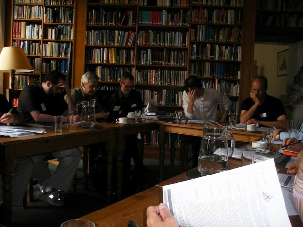 Eine Gruppe von Menschen sitzt um einen Tisch in einer Bibliothek, mit Gläsern, Tassen und Papieren auf dem Tisch und einer Lampe auf der linken Seite.