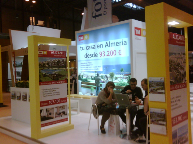 Eine Gruppe von Menschen, die um einen Tisch sitzen und auf einen Laptop schauen, mit verschiedenen Werbeplakaten daneben.