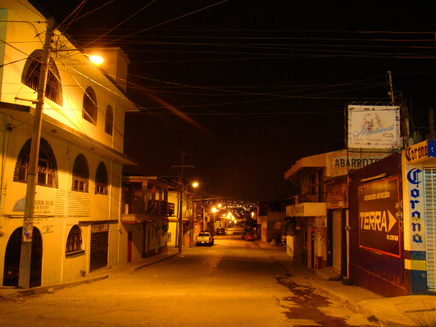 Eine Straßenszene mit Gebäuden und Strommasten auf beiden Seiten, einigen geparkten Fahrzeugen und einer Straße in der Mitte.