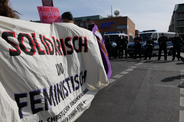 Eine Gruppe von Menschen marschiert auf einer Straße, hält ein Banner mit der Aufschrift "Solidarität und Feminismus" in den Händen, mit parkenden Fahrzeugen und Gebäuden im Hintergrund unter einem klaren blauen Himmel.