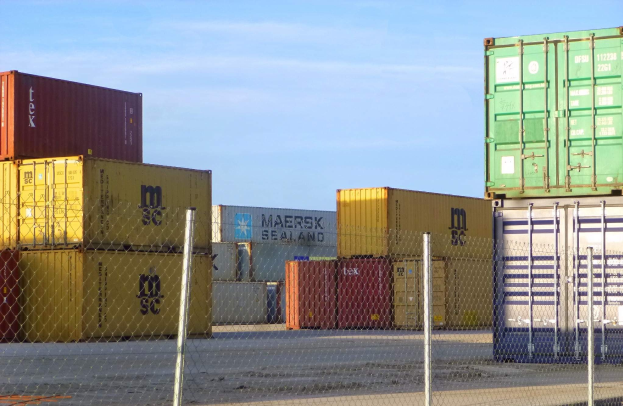 Containerstapel mit Maschendrahtzaun im Vordergrund und einer Straße darunter, unter einem bewölkten Himmel.