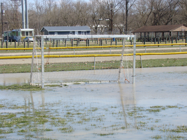 Ein Fußballtor steht in der Mitte eines überfluteten Feldes, umgeben von Gras und Wasser, vor dem Hintergrund von Schuppen, Pfählen, Bäumen, Fahrzeugen und einem klaren blauen Himmel.