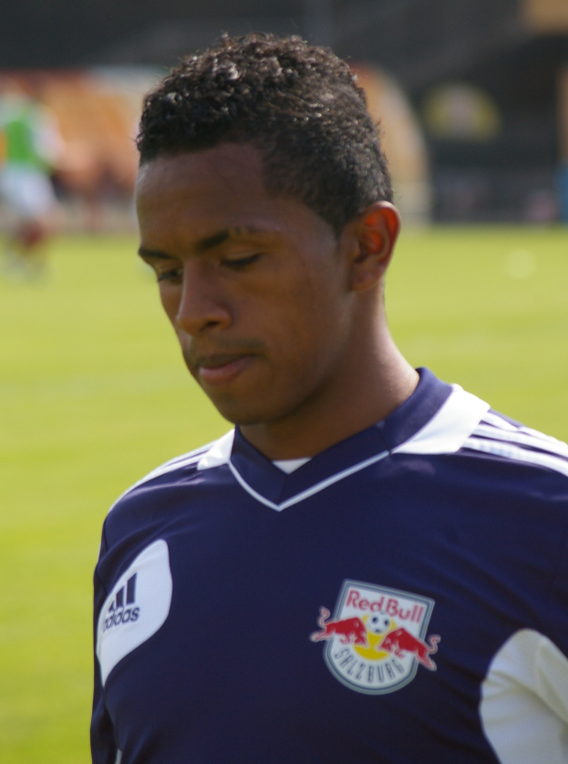 Ein junger Mann in einem blauen T-Shirt steht auf einem Fußballfeld und sieht aus wie ein Flügelstürmer mit kurzen, dunklen Haaren und einem entschlossenen Gesichtsausdruck.