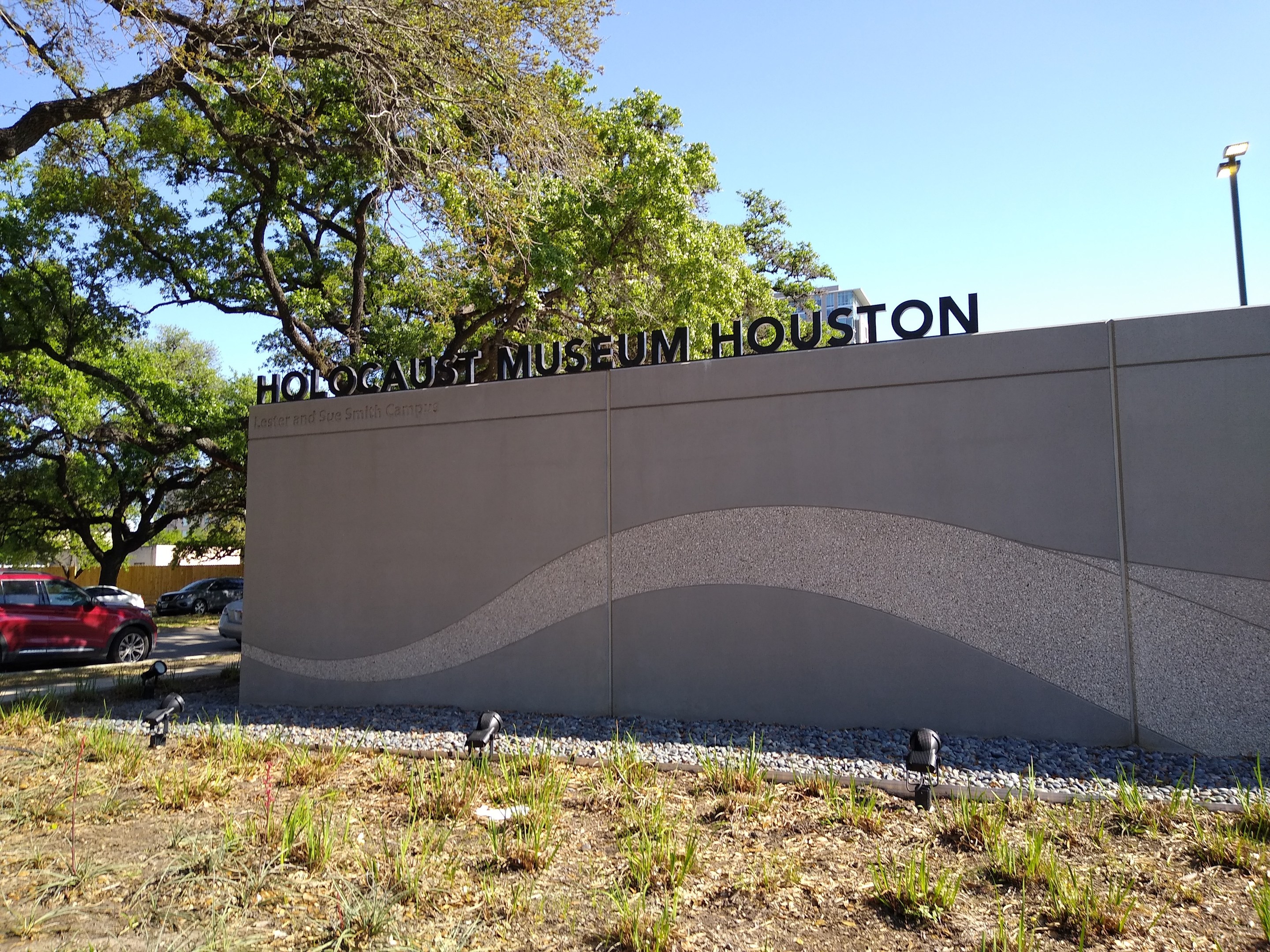 Außenansicht des Houstoner Holocaust-Museums mit eingravierter Textwand, Gedenksteine, Grünflächen, Straßenbegleitende Elemente und bewölktem Himmel.