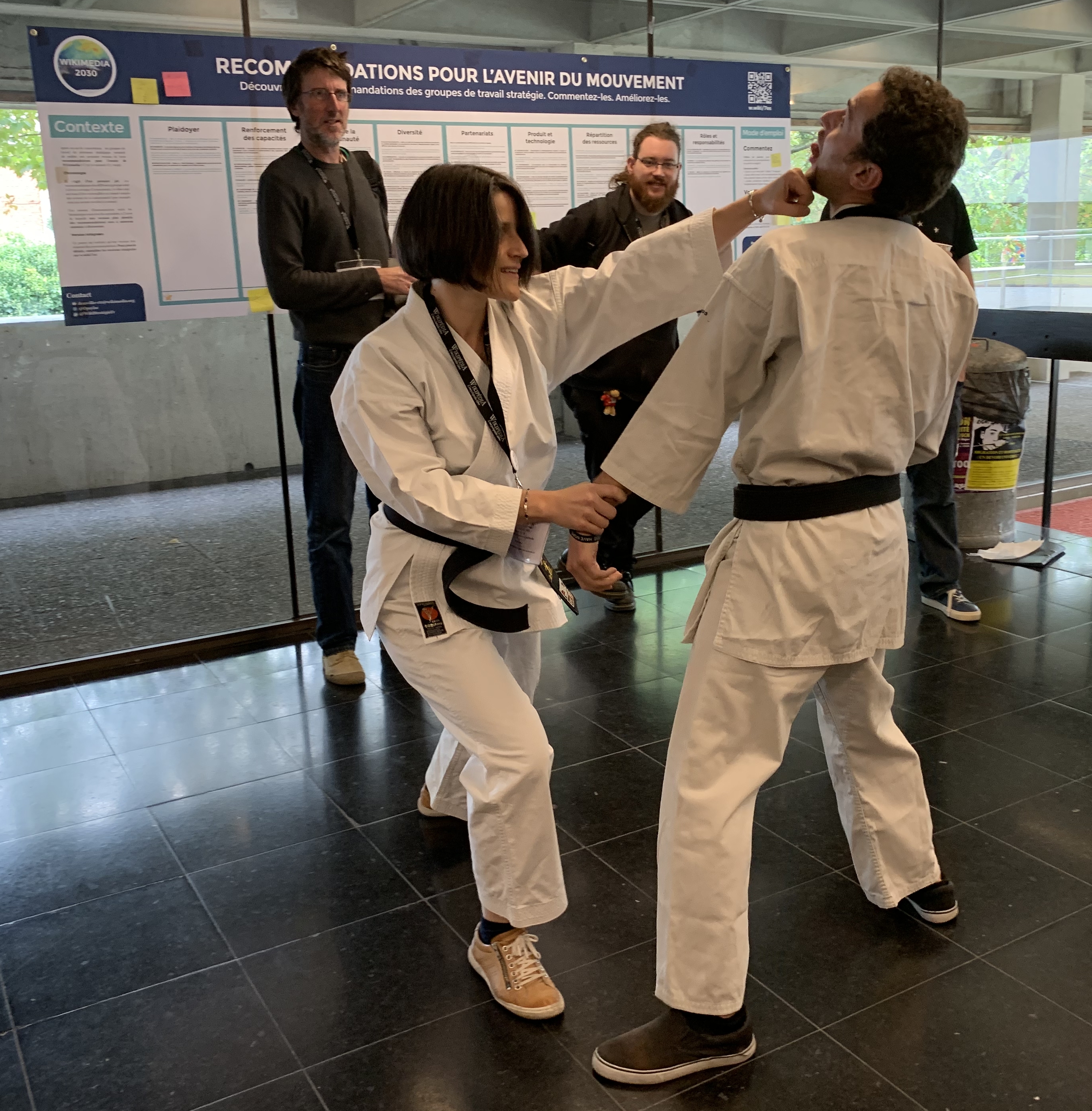 Ein Mann und eine Frau in Karate-Kleidung stehen nebeneinander vor einem Plakat, mit einer Glaswand dahinter, die Bäume zeigt, und einem Müllcontainer und einem Tisch mit Gegenständen rechts, was auf eine Taekwondo-Wettkampf-Situation hinweist.