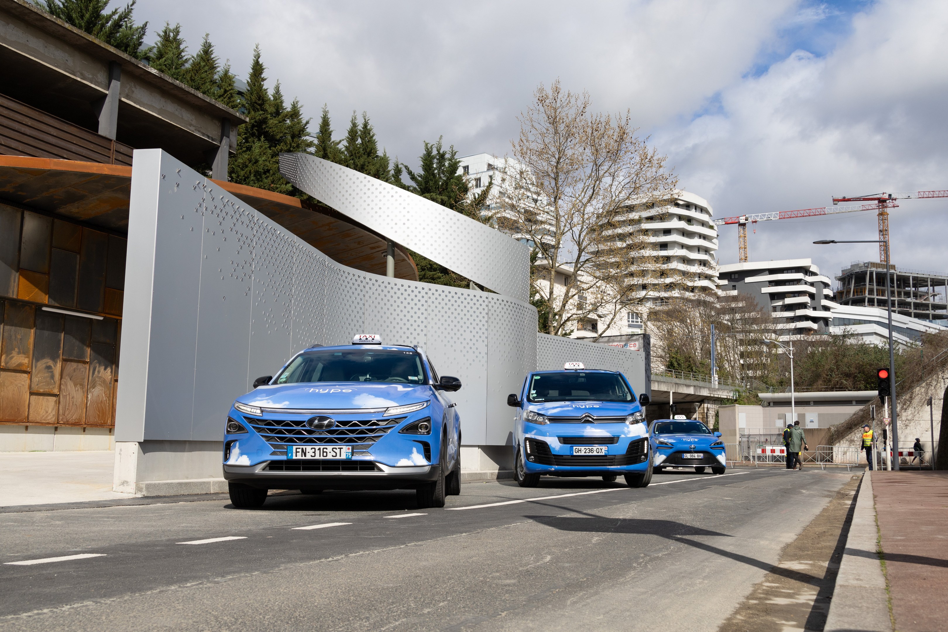 Zwei blaue autonome Autos fahren auf einer Straße neben einem Gebäude, mit Fußgängern auf dem rechten Gehweg, vor einer Kulisse aus Bäumen, Gebäuden, Kränen, Polen, Verkehrsampeln und einem klaren blauen Himmel.