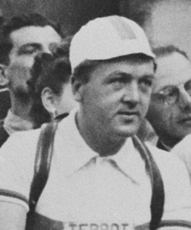 Schwarzes und weißes Foto eines Mannes mit Mütze und Hosenträgern, der vor einer Gruppe von Menschen steht, der als legendärer Fußballspieler gilt.