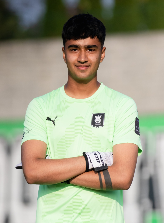 Ein junger Mann in einem grünen T-Shirt steht auf einem Fußballfeld und schaut selbstbewusst in die Kamera, mit einem unscharfen Hintergrund.