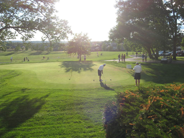 Ein Mann hält einen Golfschläger auf einem Golfplatz mit Gruppen von Menschen drumherum, Bäumen mit Zweigen und Blättern, Gras, geparkten Autos und Häusern im Hintergrund.