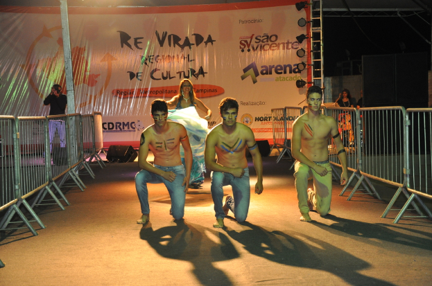 Drei shirtlose Jungen knien auf dem Boden während eines kulturellen Ereignisses, mit Grills auf beiden Seiten und einem Banner im Hintergrund, während eine Person ein Foto mit einer Kamera auf der linken Seite macht, unter Deckenbeleuchtung.