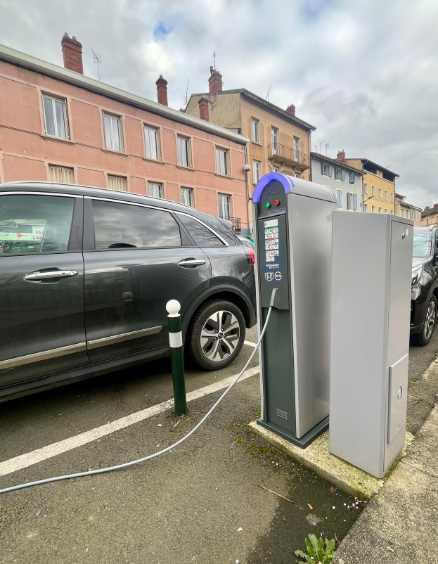Ein Elektroauto ist an einer Ladestation an der Straße angeschlossen, mit Gebäuden, Laternenpfählen und einem bewölkten Himmel im Hintergrund.
