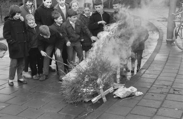 Gruppe von Kindern, die um ein Feuer auf einem Gehweg stehen, einige halten Stöcke, mit Fahrrädern, Pfählen, Bäumen und Gebäuden im Hintergrund in Schwarz-Weiß.
